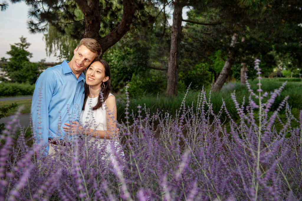 GPT
A couple embraces amid a field of purple flowers, with evergreen trees and a serene park-like setting in the background. The man, wearing a light blue shirt, gently wraps his arms around the woman, who is dressed in a white sleeveless top, smiling comfortably as she leans back into him. The warm, natural setting gives a sense of tranquility and romantic connection.
