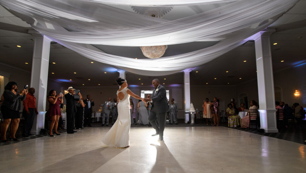 A couple sharing their first dance as newlyweds in a spacious reception hall adorned with white drapes and a large chandelier.
