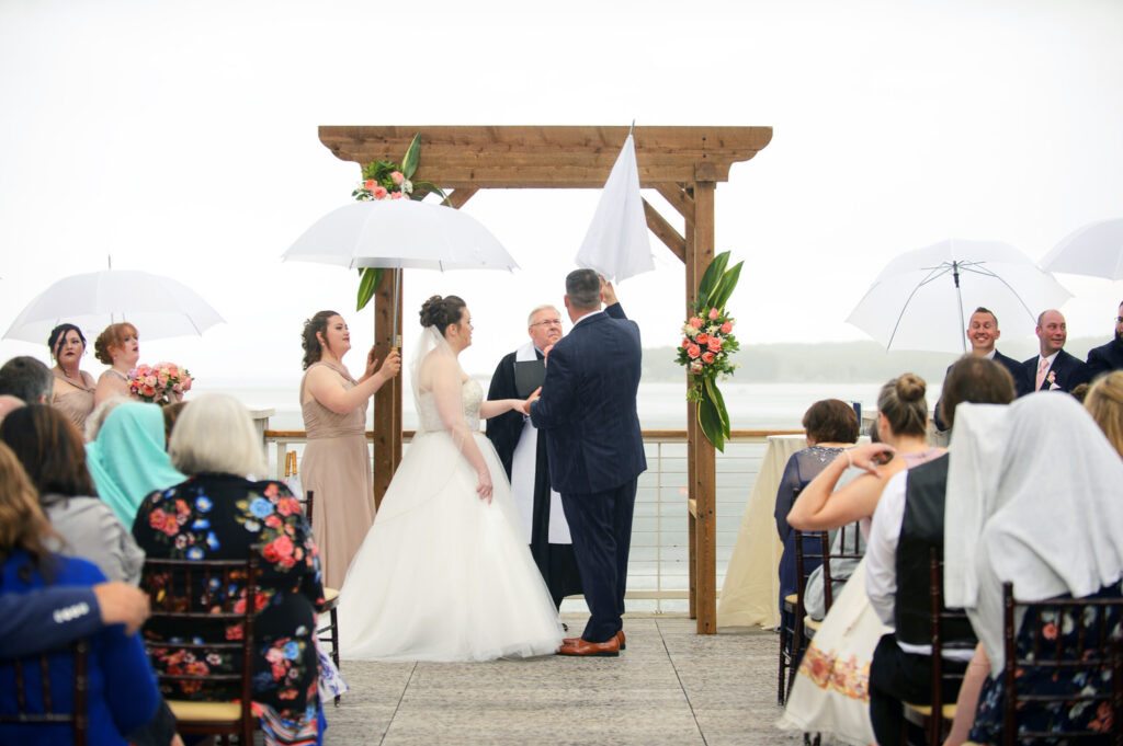 An outdoor wedding ceremony under gray skies with guests holding umbrellas and a smiling bridal party by a wooden altar with lake views.
