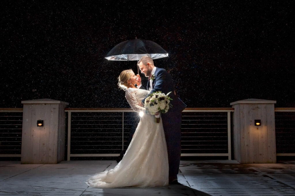A newlywed couple sharing a romantic kiss under an umbrella on a rainy evening, with soft lighting creating a dramatic and intimate atmosphere.