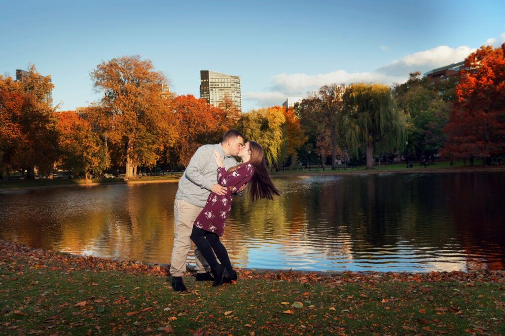 n this vibrant autumn scene, a couple is mid-dance on the leaf-strewn grass of Boston Common, sharing a kiss. The woman, dressed in a maroon floral dress, is swept off her feet by a man in a grey sweater, against a backdrop of colorful fall foliage and a serene pond reflecting the warm glow of the setting sun.