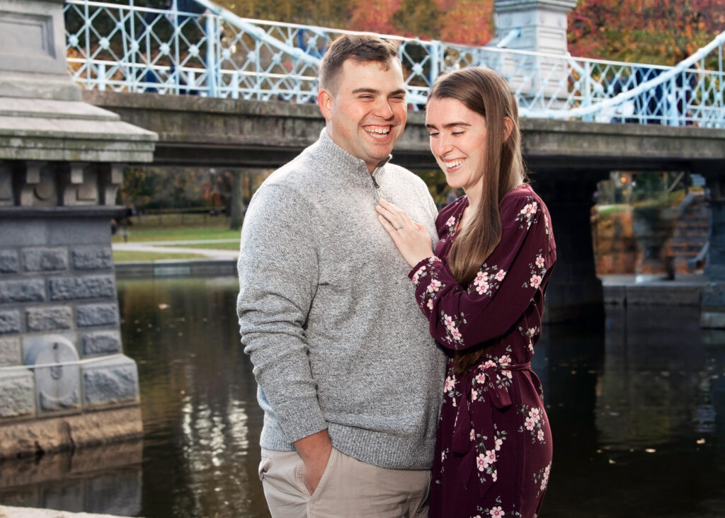 Engaged couple standing on a bridge at Public Garden, Boston, with colorful fall foliage and the blue bridge in the background