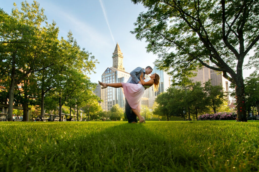 GPT
In a lush park with the Boston skyline as a backdrop, a couple shares a dip kiss, captured from a low angle that accentuates the vibrant green grass and the city's architecture. The woman, wearing a pink flowing skirt, is lovingly cradled in the man's arms as they share a romantic moment, with the historic Custom House Tower peeking through the trees.