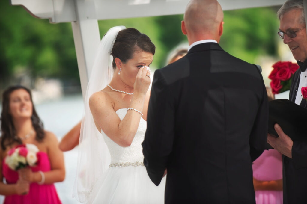 A touching moment as the bride, in a beautifully detailed wedding gown, wipes a tear from her eye during the ceremony, with an out-of-focus backdrop of the groom and wedding guests