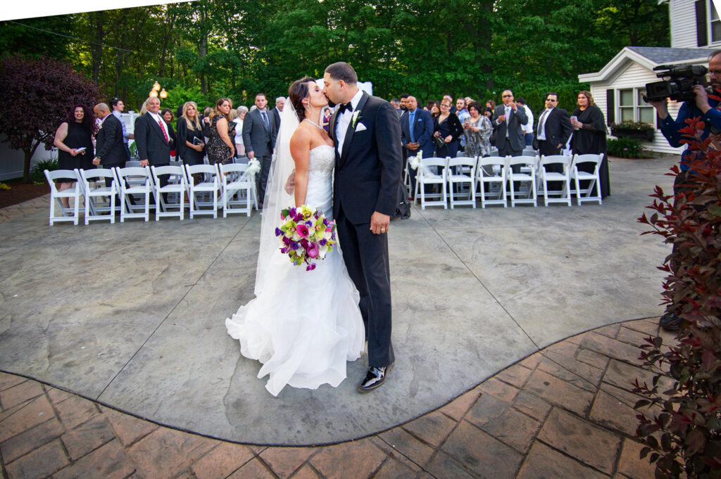Bride and groom kissing in the center of a paved courtyard, with guests watching on and a videographer capturing the moment, signifying the joyous culmination of their wedding ceremony