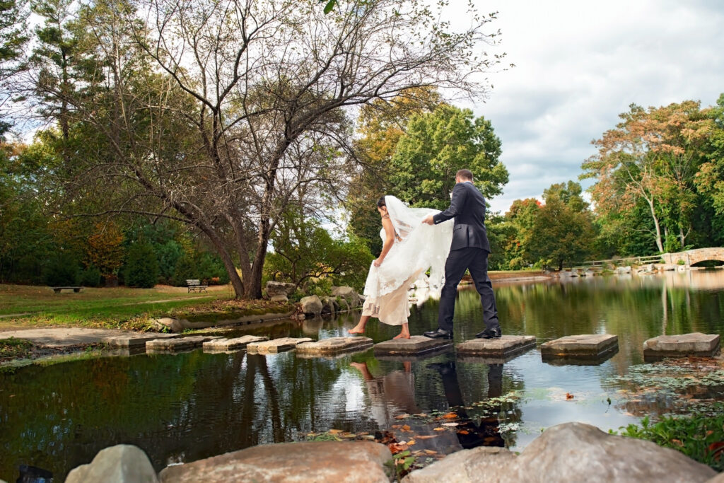 A bride and groom carefully traverse stepping stones across a serene pond, surrounded by the lush greenery of a park, capturing a moment of adventurous spirit on their wedding day