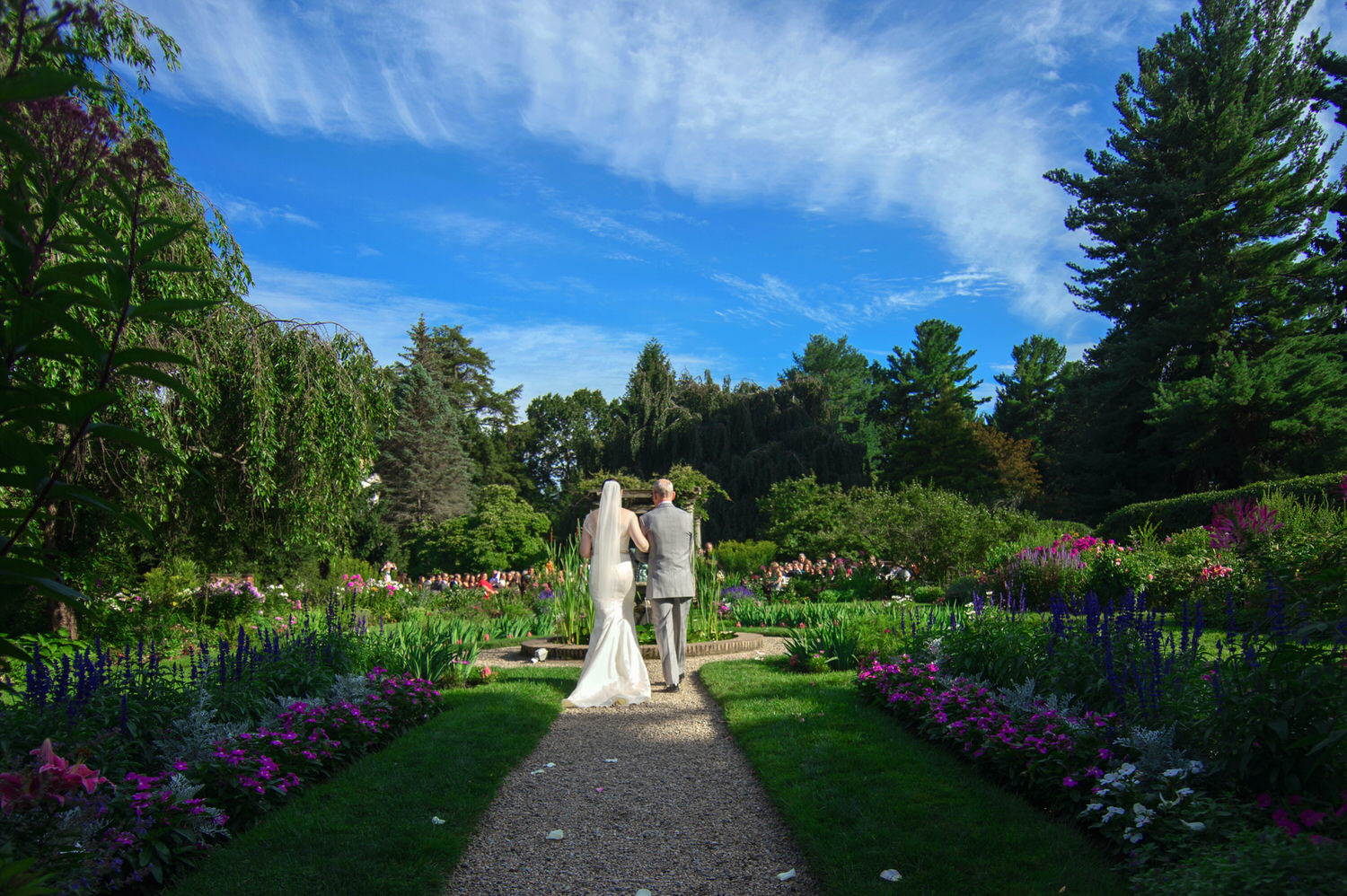 A bride and groom walking hand in hand along a garden path with vibrant flowers and clear blue skies above, depicting a serene outdoor wedding setting at one of the best wedding venues in Massachusetts