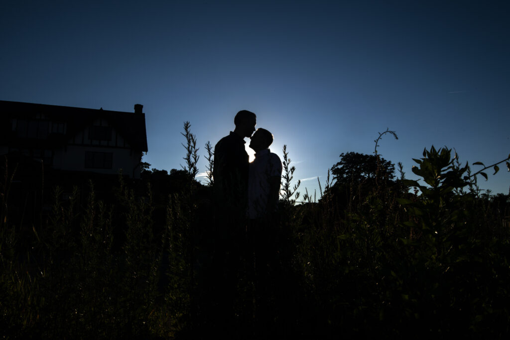 A silhouette of a couple sharing an intimate moment is captured against the striking backdrop of a luminous blue sky. The sun, sitting just behind them, creates a radiant outline highlighting their profiles. They stand amidst tall grasses, with the distinctive outline of a traditional house faintly visible in the background, evoking a serene and romantic atmosphere.