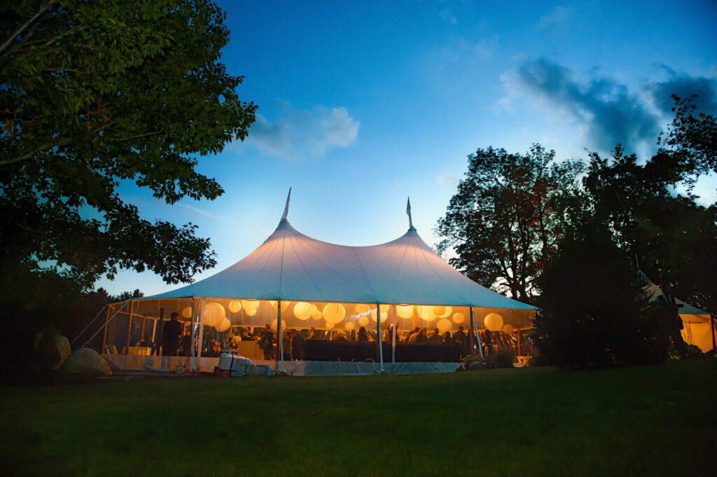 A majestic white event tent illuminated from within at twilight, nestled among trees, providing a cozy and festive atmosphere for an outdoor wedding reception.