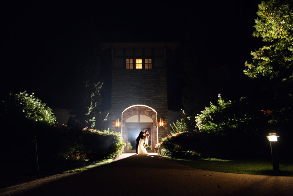 A bride and groom are silhouetted in a romantic dip under the archway of a stone building, illuminated by ambient light that sets an intimate mood against the night sky