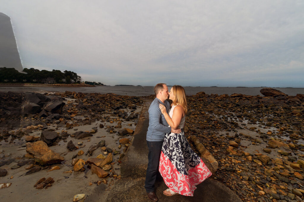 A couple stands close together on a rocky shoreline at Lynch Park in Beverly, MA, sharing an intimate moment with the serene ocean and a hazy sky in the background. The woman's dress with a bold black and red floral pattern flows in contrast to the rugged terrain.