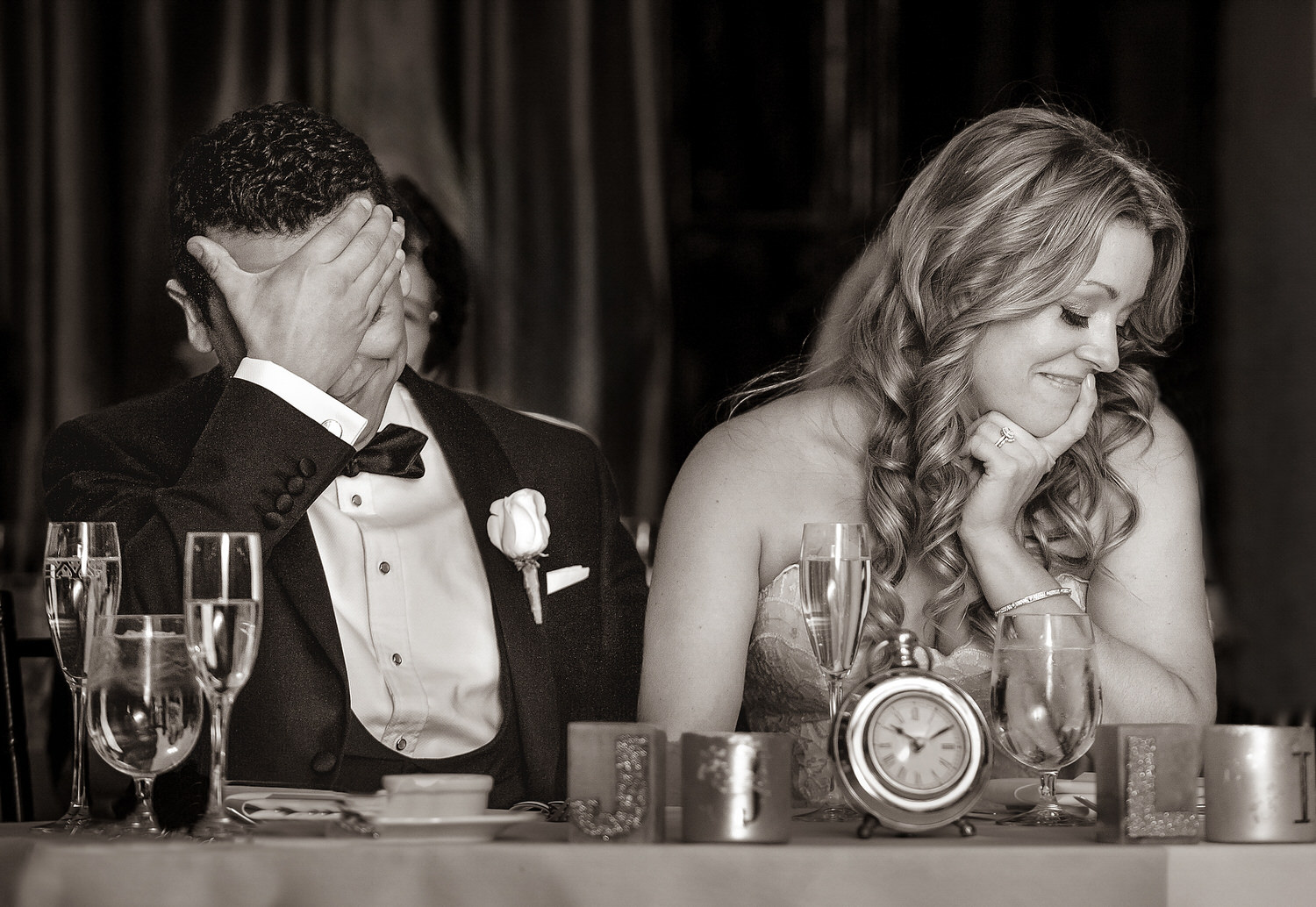 A groom facepalming and a bride smiling thoughtfully, both elegantly dressed, seated at a wedding table with a romantic setting in sepia tones in documentary style wedding photography