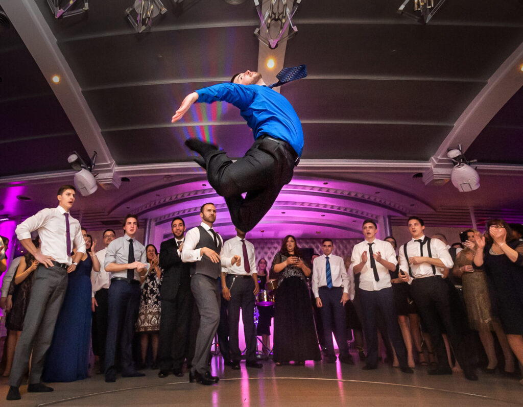 Guests circling around dance floor while man in blue shirt and black pants jumps in the air with purple lights in the background