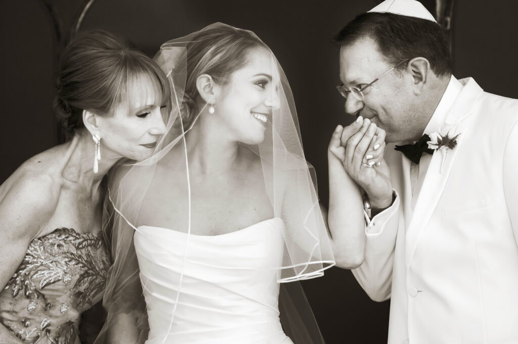 In a timeless black and white photo, a radiant bride smiles under her veil, flanked by an elegantly dressed woman on one side and a man in a white suit with a kippah, who appears moved, on the other