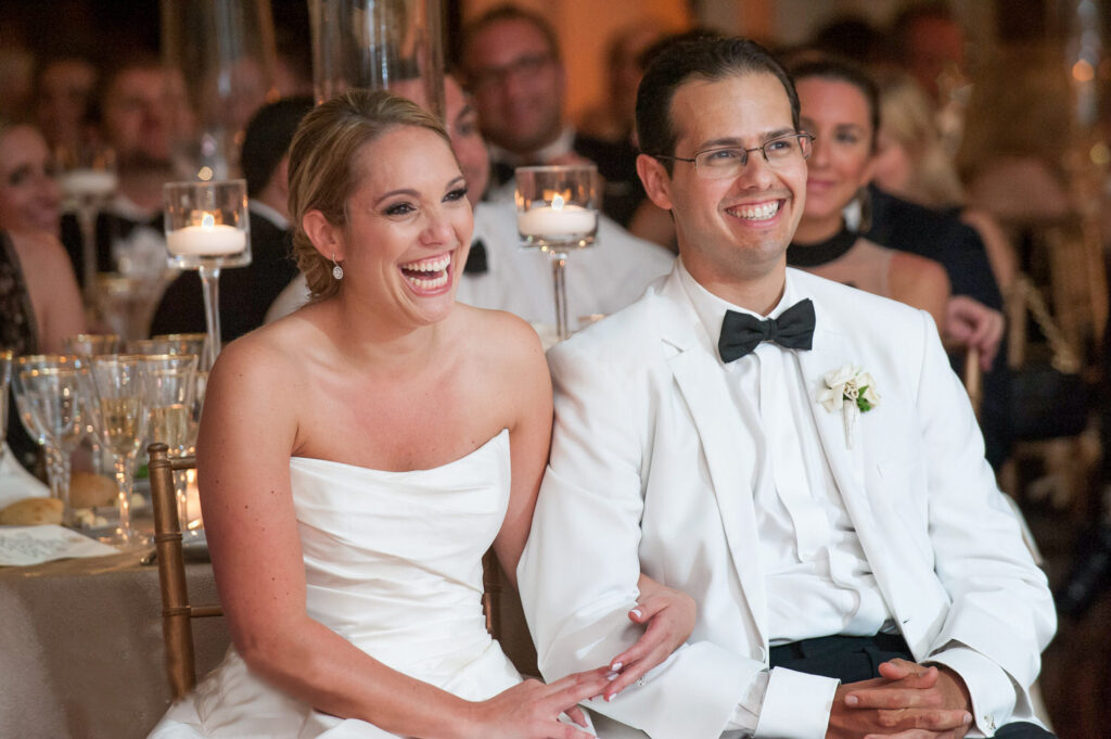 A laughing bride and a smiling groom seated at a wedding reception, enjoying the moment, surrounded by guests and candlelight ambiance