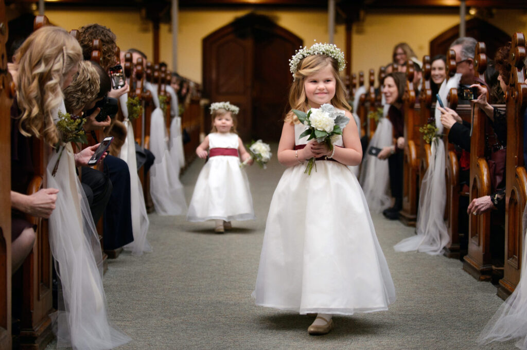 Young flower girl in white dress holding bouquet walks down aisle with another flower girl in a matching dress walking behind her