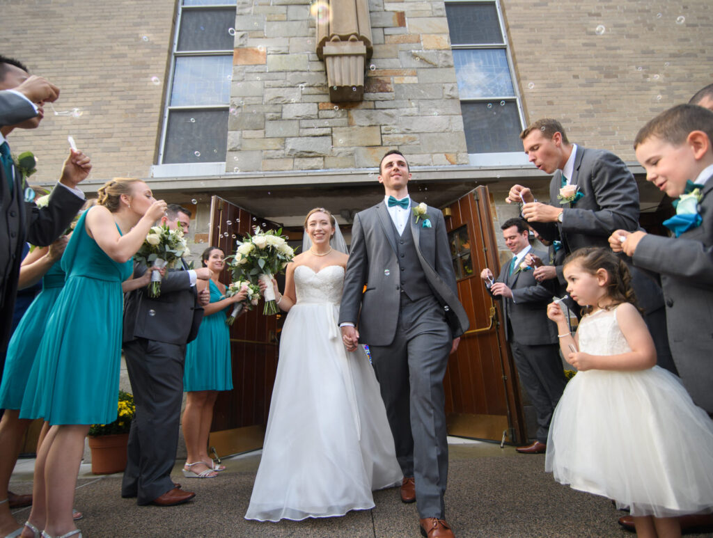 A jubilant bride and groom walk hand in hand, exiting a stone church as friends and family surrounding them blow bubbles in celebration, with smiles and festive attire completing the scene