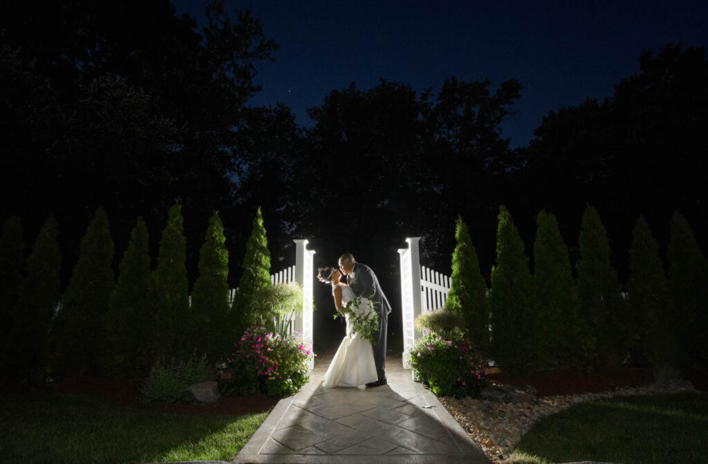 A couple shares a kiss on a lit garden path at night, creating a romantic scene with the light casting a glow around their silhouette.