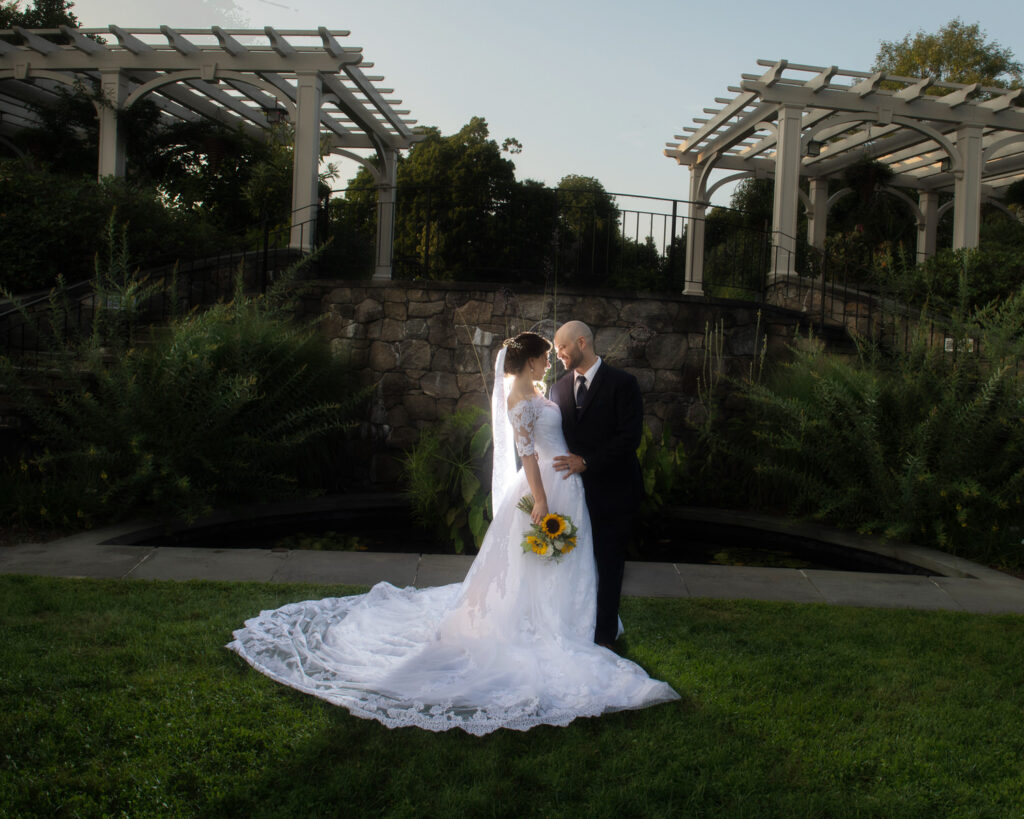 Bride and groom in an embrace at the Tower Hill Botanical Garden, with architectural elements and lush plants framing the serene moment.