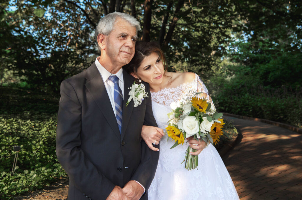 Bride holding a bouquet of sunflowers leaning on her father's shoulder, both smiling, before a lush green backdrop.