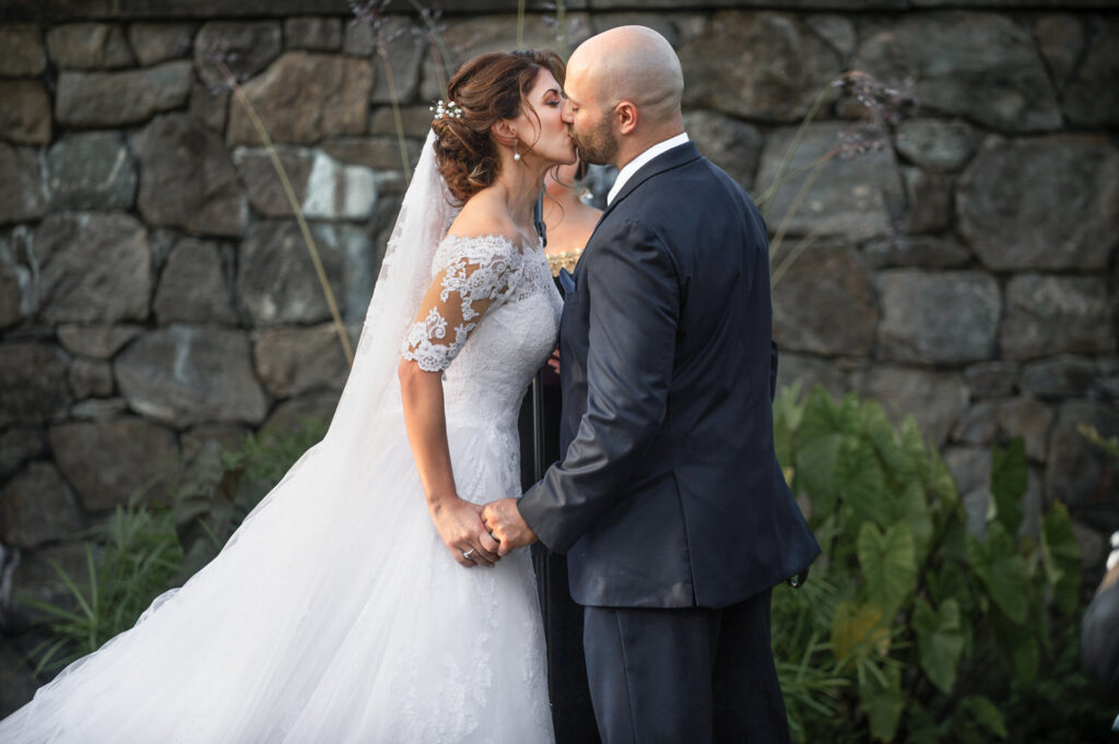 Newlyweds share a romantic kiss in front of a stone wall, surrounded by greenery, in the peaceful setting of a botanical garden.