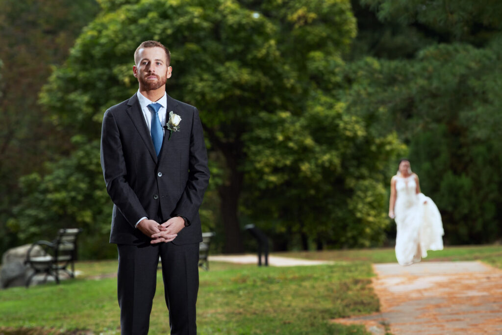 Groom standing in green park with bride in background holding her gown train in her hand