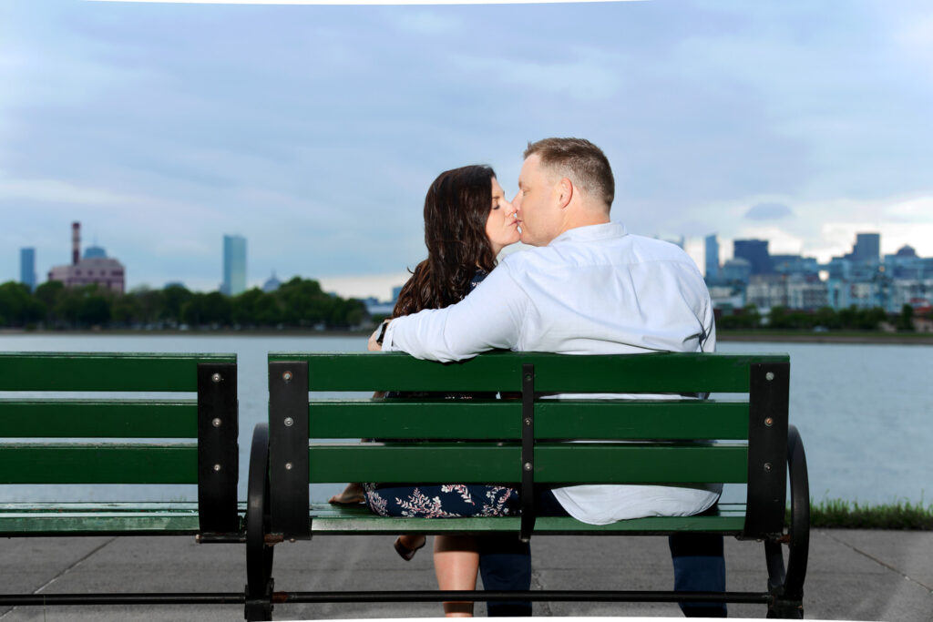 A couple sits closely on a green park bench, sharing a kiss on the cheek while overlooking a calm body of water with a cityscape in the distance. The man, dressed in a white long-sleeve shirt, has his back to the camera, while the woman, wearing a patterned dress and white sandals, faces the camera. The setting is tranquil, with the backdrop featuring an overcast sky and urban buildings, hinting at a romantic moment in an urban park setting.