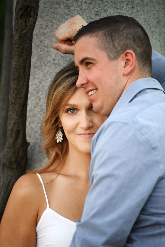 Close-up of a smiling couple in casual attire, leaning against a granite wall, engaged in a tender moment
