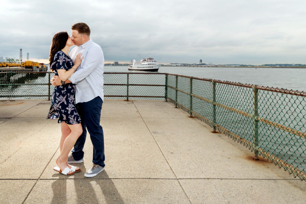 A couple shares an intimate embrace on Castle Island overlooking Boston Harbor, with a passenger ferry and the distant city skyline in the background. The cloudy sky suggests a mood of calm anticipation, adding depth to the romantic scene.
