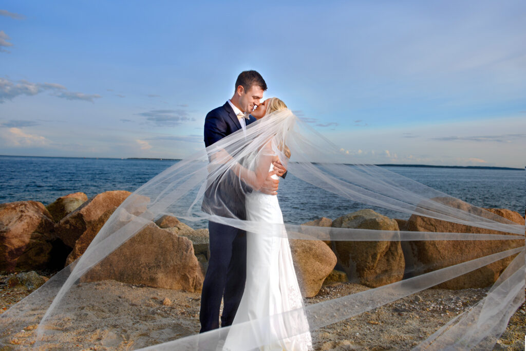 Newlyweds embracing on a sandy beach, the bride's veil caught in a gust, creating a translucent shroud around them, with the sea and sky as a tranquil backdrop