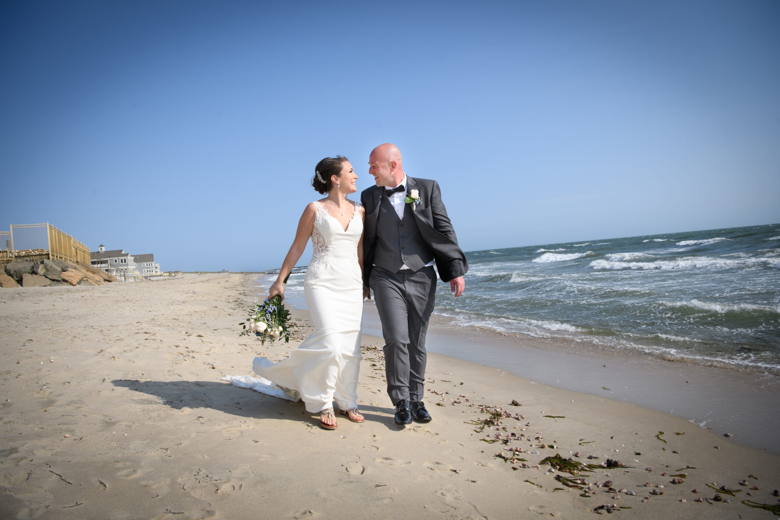 Bride in white lace dress and groom in suit walk down the beach on a sunny day highlighting a wedding timeline with first look