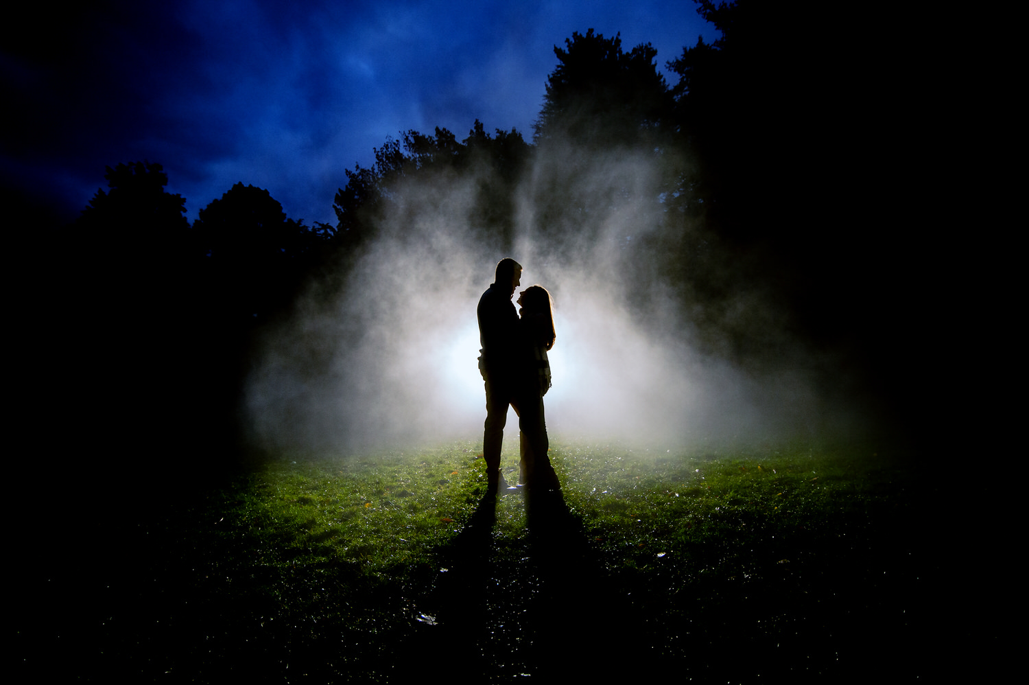 GPT Silhouetted couple embracing in a misty field at the Arnold Arboretum in Jamaica Plain, with a dramatic backlight casting a mystical glow and creating a serene, romantic atmosphere showing when to take engagement photos.