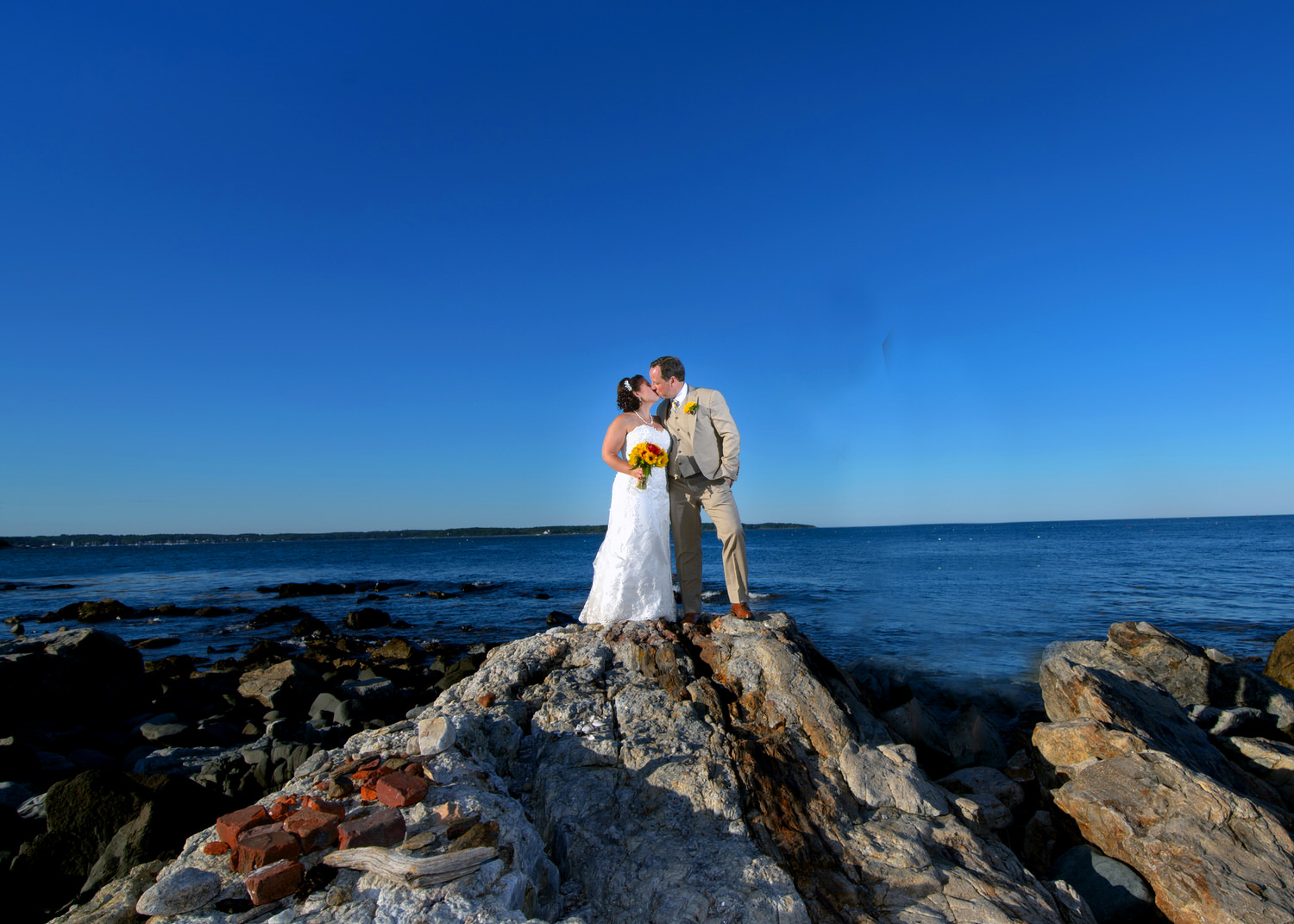 Bride and groom sharing a kiss on a rocky shore with the ocean stretching into the horizon under a clear blue sky showing wedding photography tips for couples