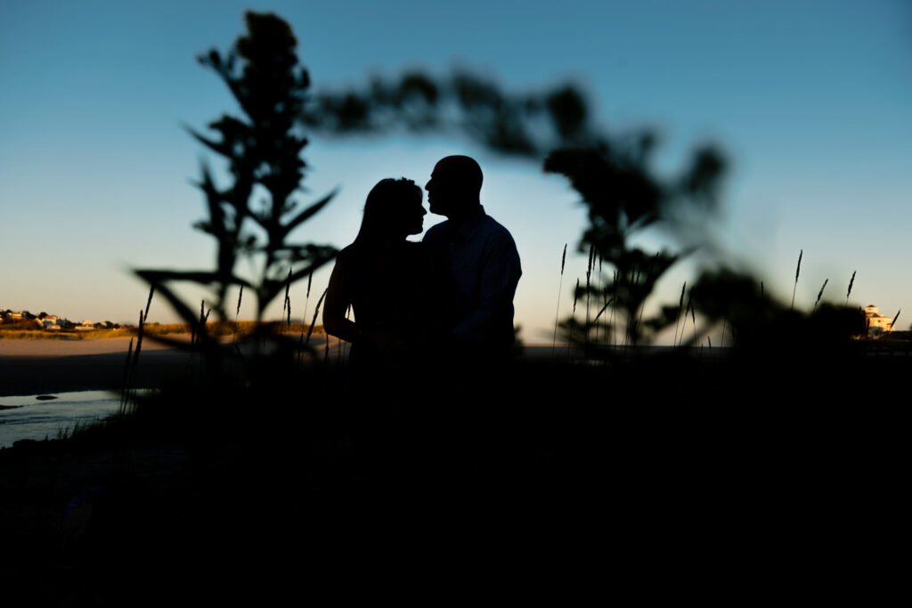 A silhouette of a couple standing close together, their profiles outlined by the golden hues of a sunset at Good Harbor Beach in Gloucester, MA. Wild grasses in the foreground add depth to the scene, with the calm coastal landscape stretching into the distance.