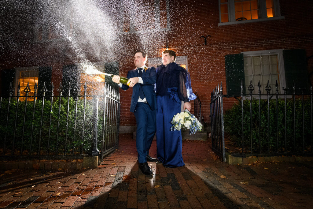 A celebratory moment outside the Hamilton House with a couple popping a champagne bottle; the cork and bubbles flying, as they smile joyfully, clad in elegant wedding attire against the historical brick backdrop.