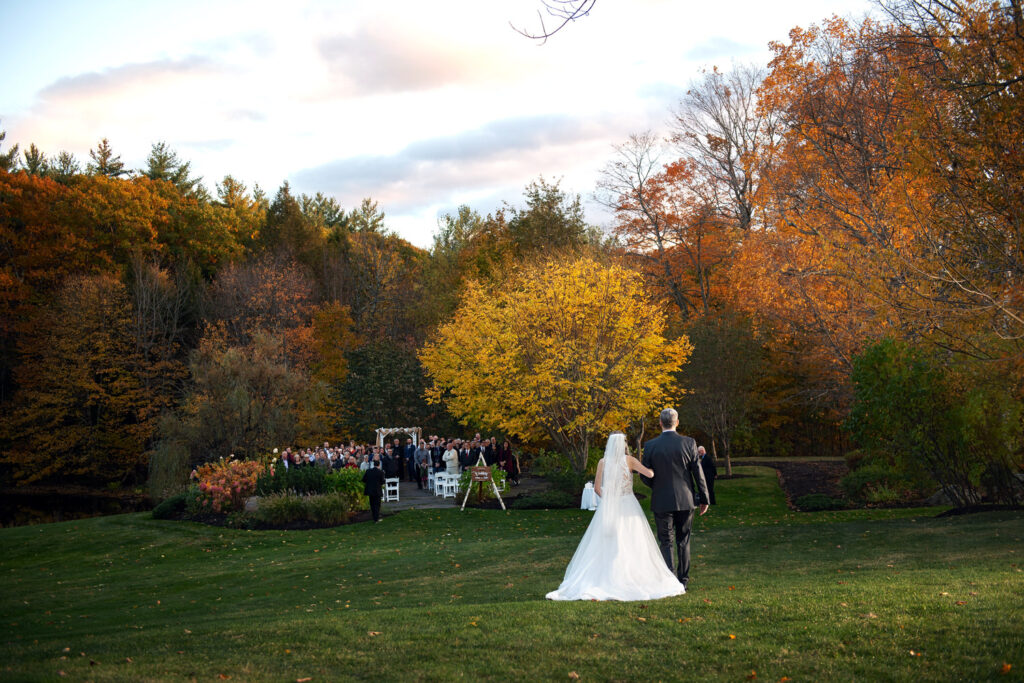 A bride and groom walking hand in hand towards an outdoor wedding ceremony with guests seated by an autumnal forest backdrop at dusk.