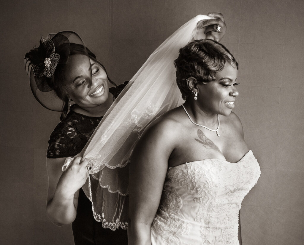 Black and white photo of bride having veil put on by smiling woman