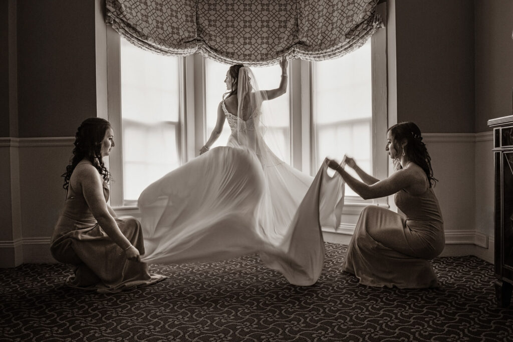 Black and white photo of a bride in her wedding dress caught in a moment of joy as two bridesmaids help fluff the train by a window.