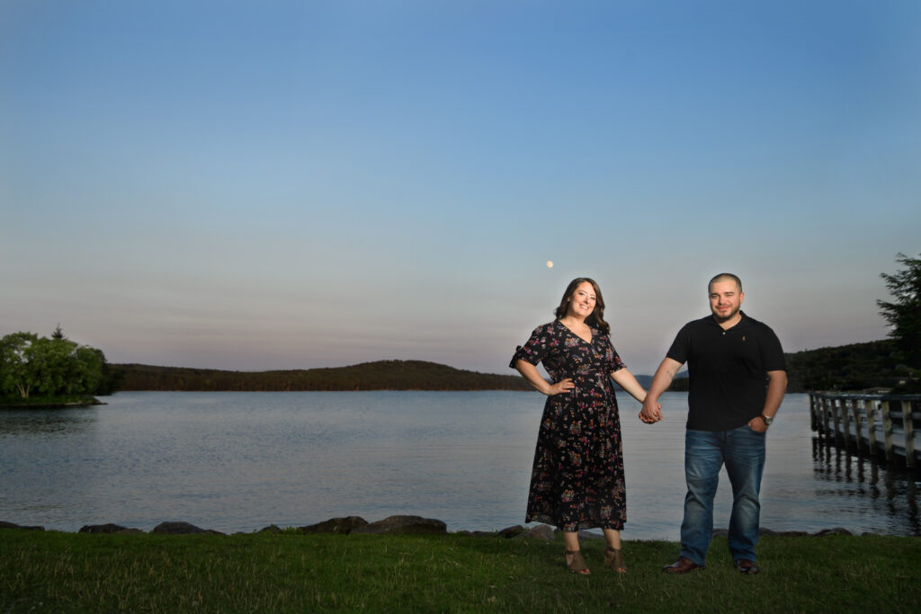 Standing on the lush green shore of a tranquil lake in New Hampshire's White Mountains, a couple holds hands and smiles towards the camera. She's dressed in a floral midi dress, and he's in a casual black polo and jeans. The soft glow of twilight illuminates their faces, and a gentle moonrise adds a touch of romance to the serene landscape, with forested hills rolling softly into the distance.