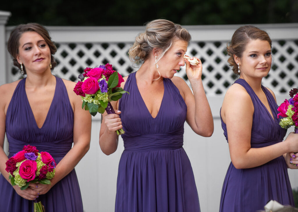 Three bridesmaids in purple dresses, one wiping away a tear, holding vibrant pink and purple bouquets at a wedding ceremony.
