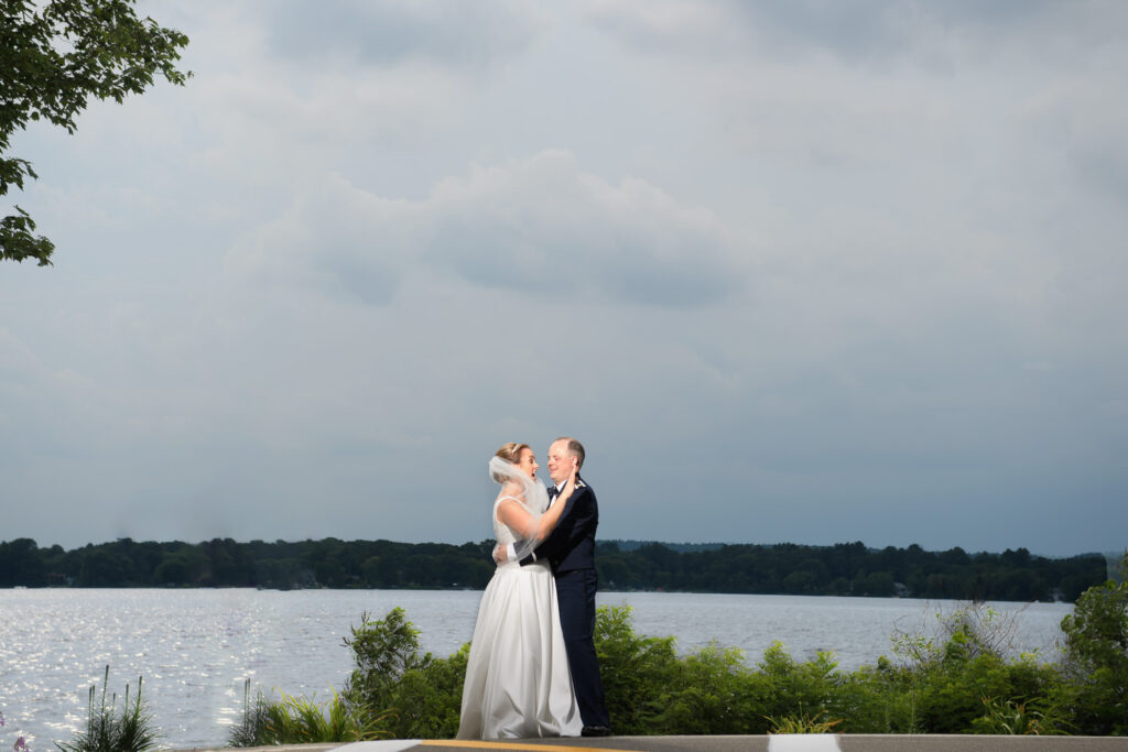 A bride and groom share an embrace on an elevated shoreline overlooking a glistening lake, with overcast skies above that promise a dramatic backdrop to their tender moment.