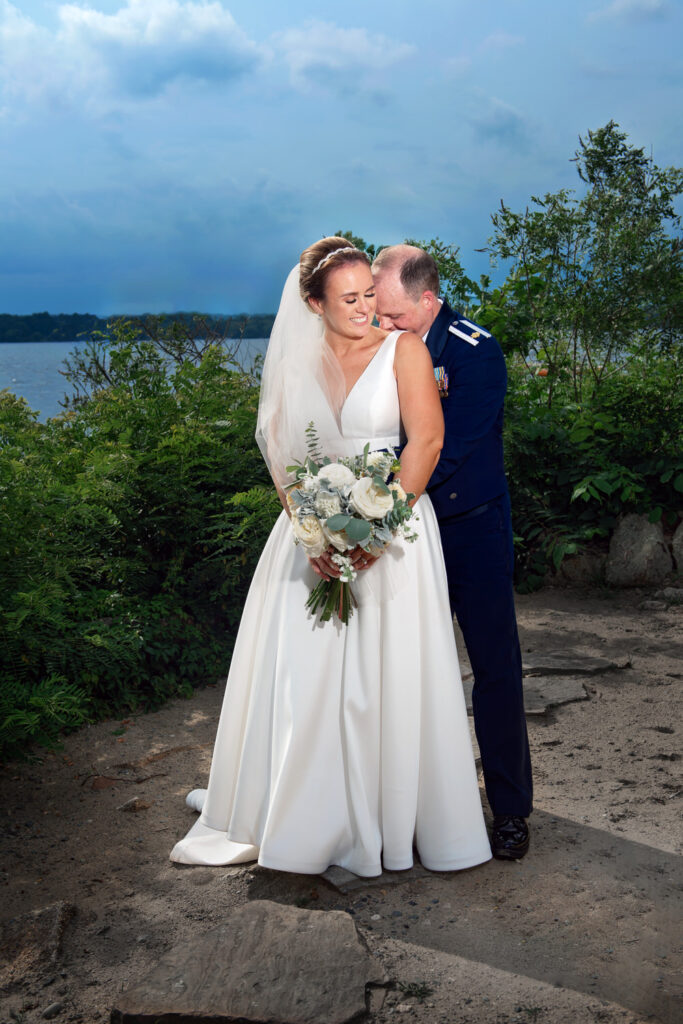 Bride in a white dress and a military groom in dress uniform sharing a quiet moment by a lakeside, with stormy skies in the background.