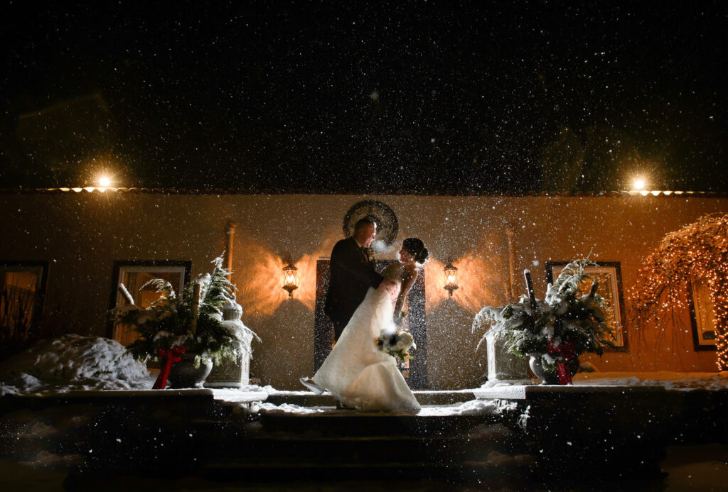 A bride and groom share an enchanting kiss on a snowy evening, framed by festive holiday decorations and warmly lit architecture, with snowflakes gently falling around them, creating a magical winter wedding scene