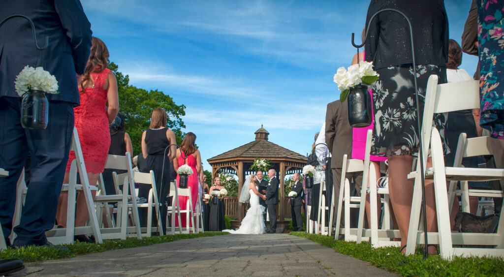 GPT A wedding ceremony at Zukas Hilltop Barn in Spencer, MA, with guests standing as the bride and groom meet at the altar, white chairs adorned with mason jars and hydrangeas lining the aisle, and a gazebo in the background under a clear blue sky.