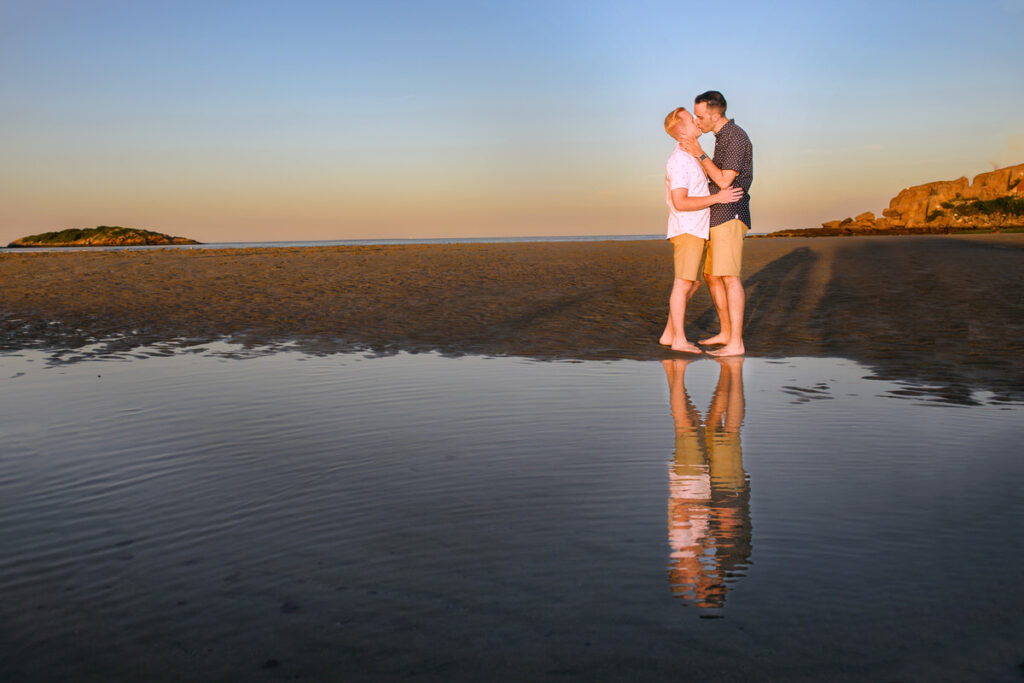 A couple kisses tenderly on a sandy beach at sunset, their reflection visible in the wet sand, creating a romantic and serene scene with an island in the background.