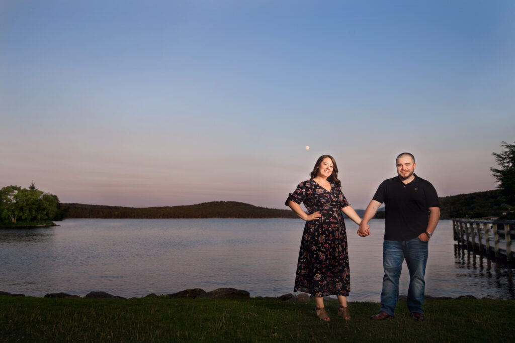 At sunset by a lake, a couple stands hand-in-hand, the woman in a floral dress smiling confidently at the camera, and the man in a casual outfit looking at her, with tranquil water and a forested hill in the background.