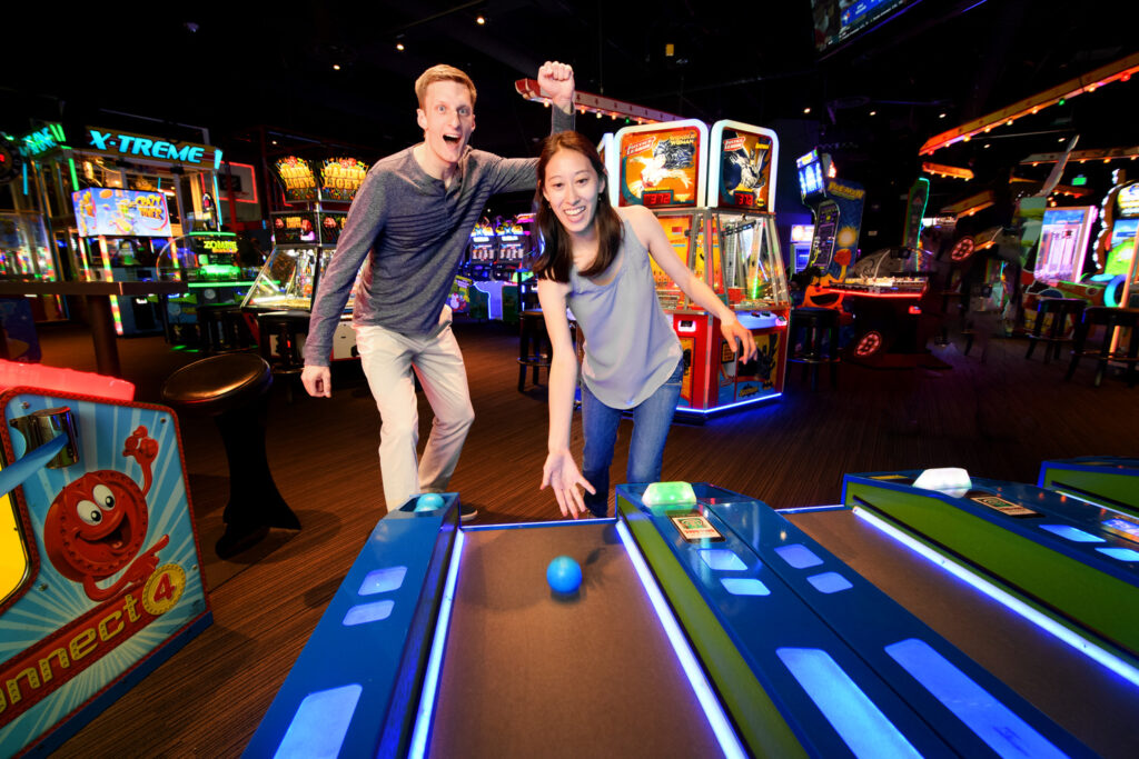 A couple excitedly plays a ball rolling arcade game in a vibrant, colorful arcade room, with the woman smiling and pushing a ball down the lane as the man celebrates beside her.