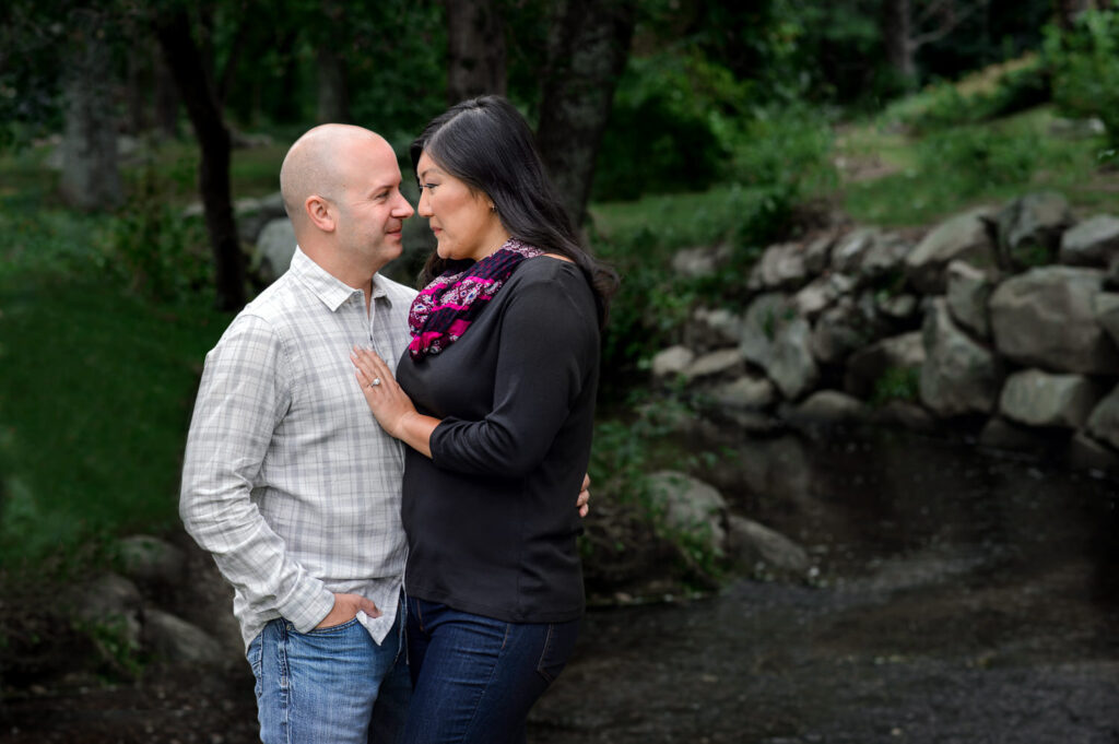 A couple stands closely together by a serene stream in a lush, green park, exchanging affectionate looks. The woman, wearing a black top and a colorful scarf, places her hand on the man's chest, who is dressed in a casual plaid shirt.
