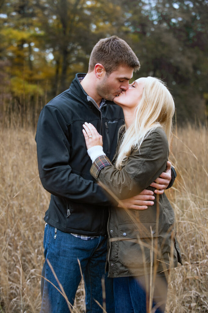 In a rustic autumn setting, a couple shares a passionate kiss among tall, golden grass with trees in soft focus in the background, symbolizing warmth and intimacy.
