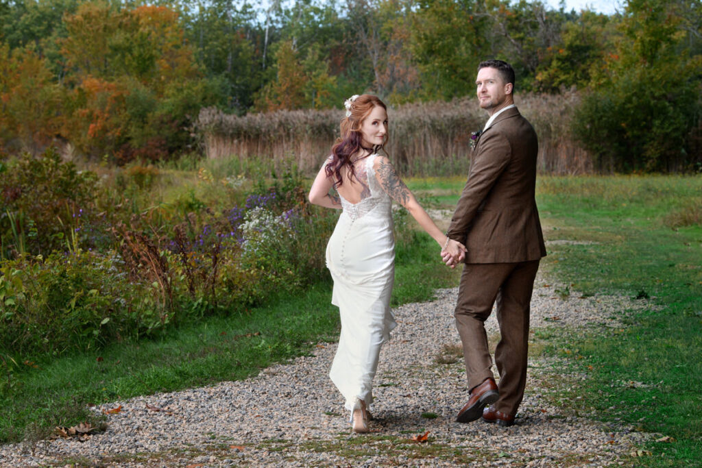 A newlywed couple holding hands and walking along a gravel path while looking back over their shoulders. 